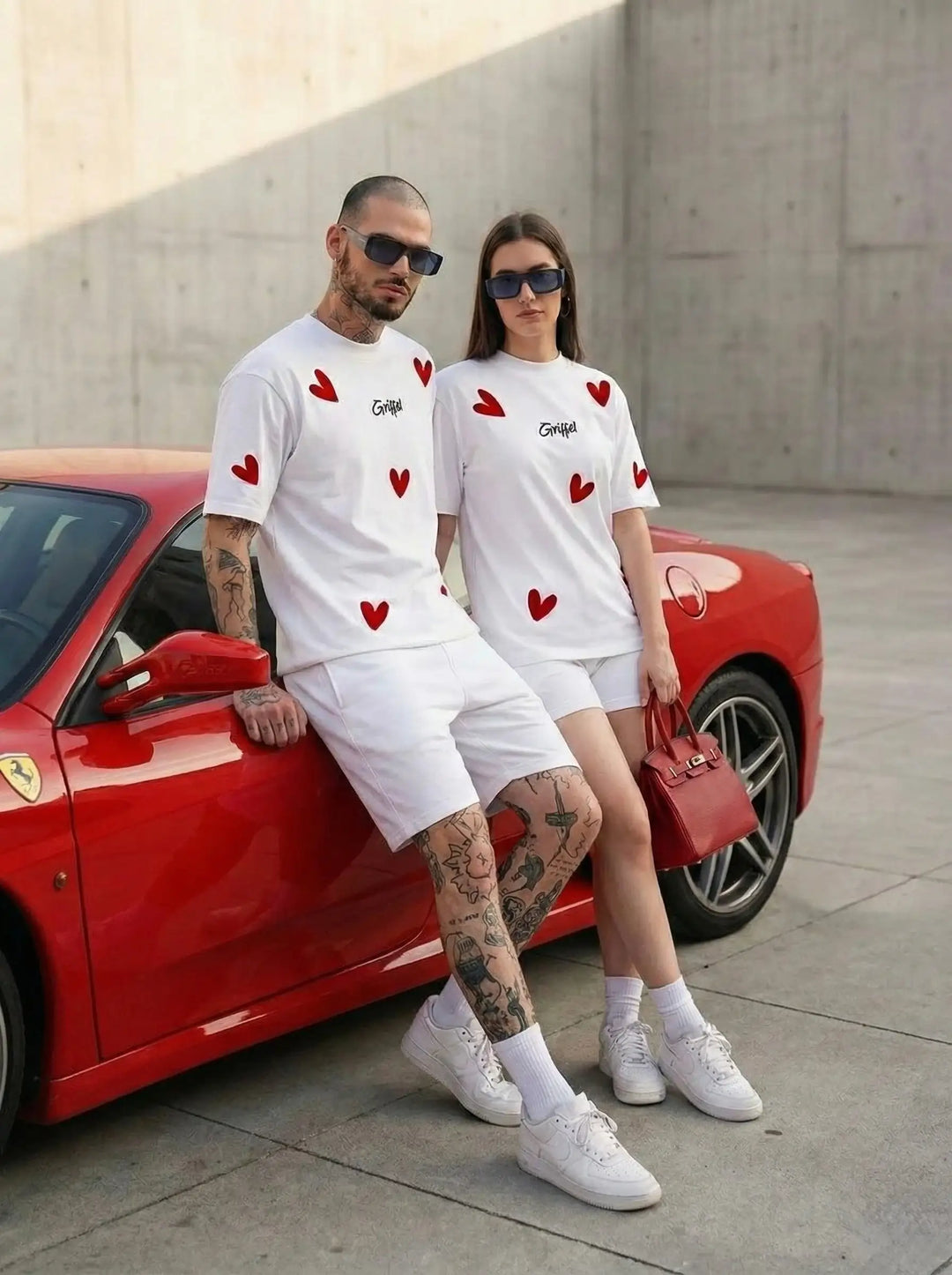 Couple in matching white heart print t-shirts and shorts, standing by a red sports car