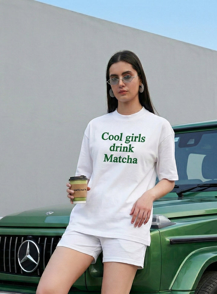 Woman in a white t-shirt with text standing next to a green Mercedes-Benz vehicle.