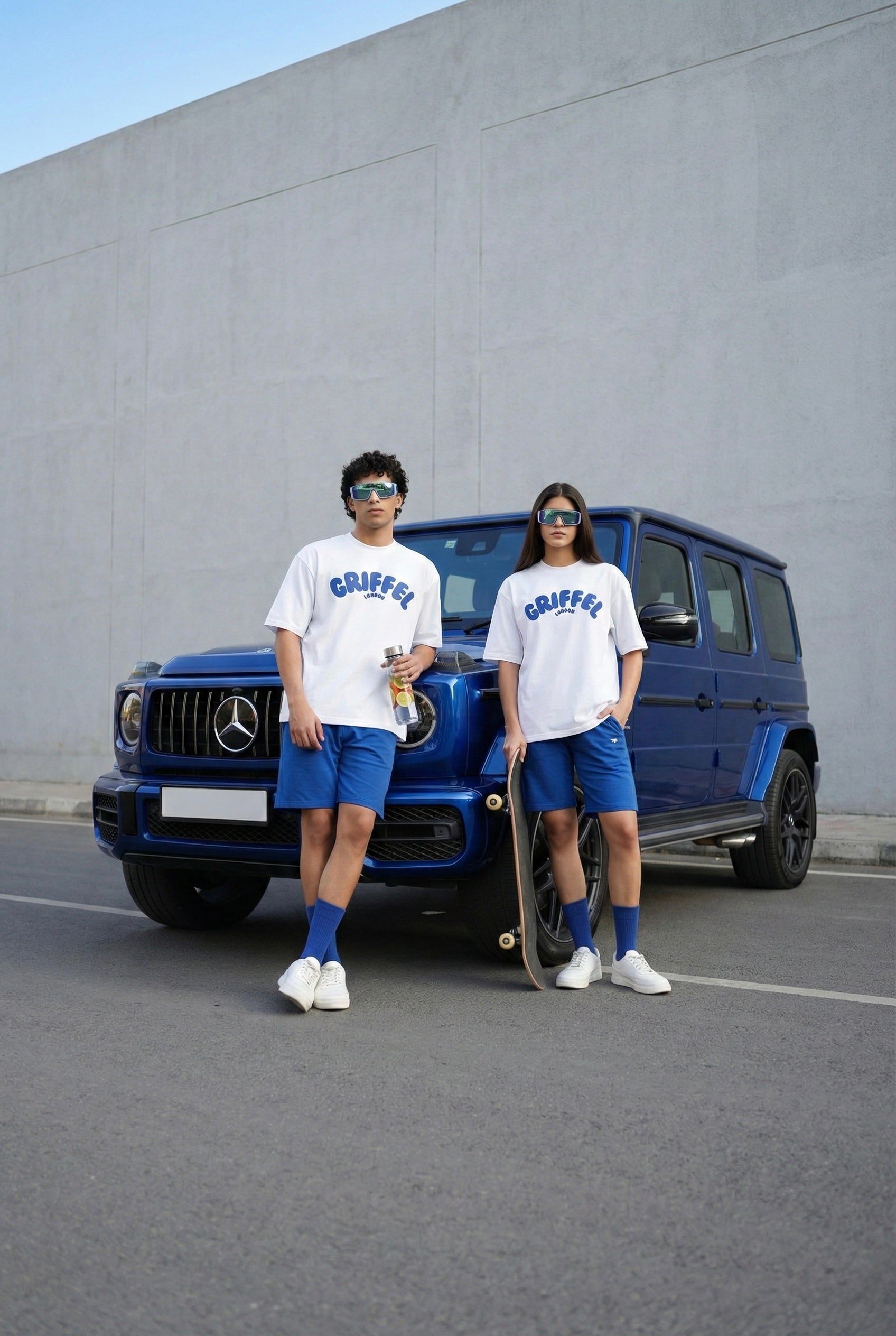 Two people in matching outfits standing next to a blue Mercedes-Benz G-Class SUV.