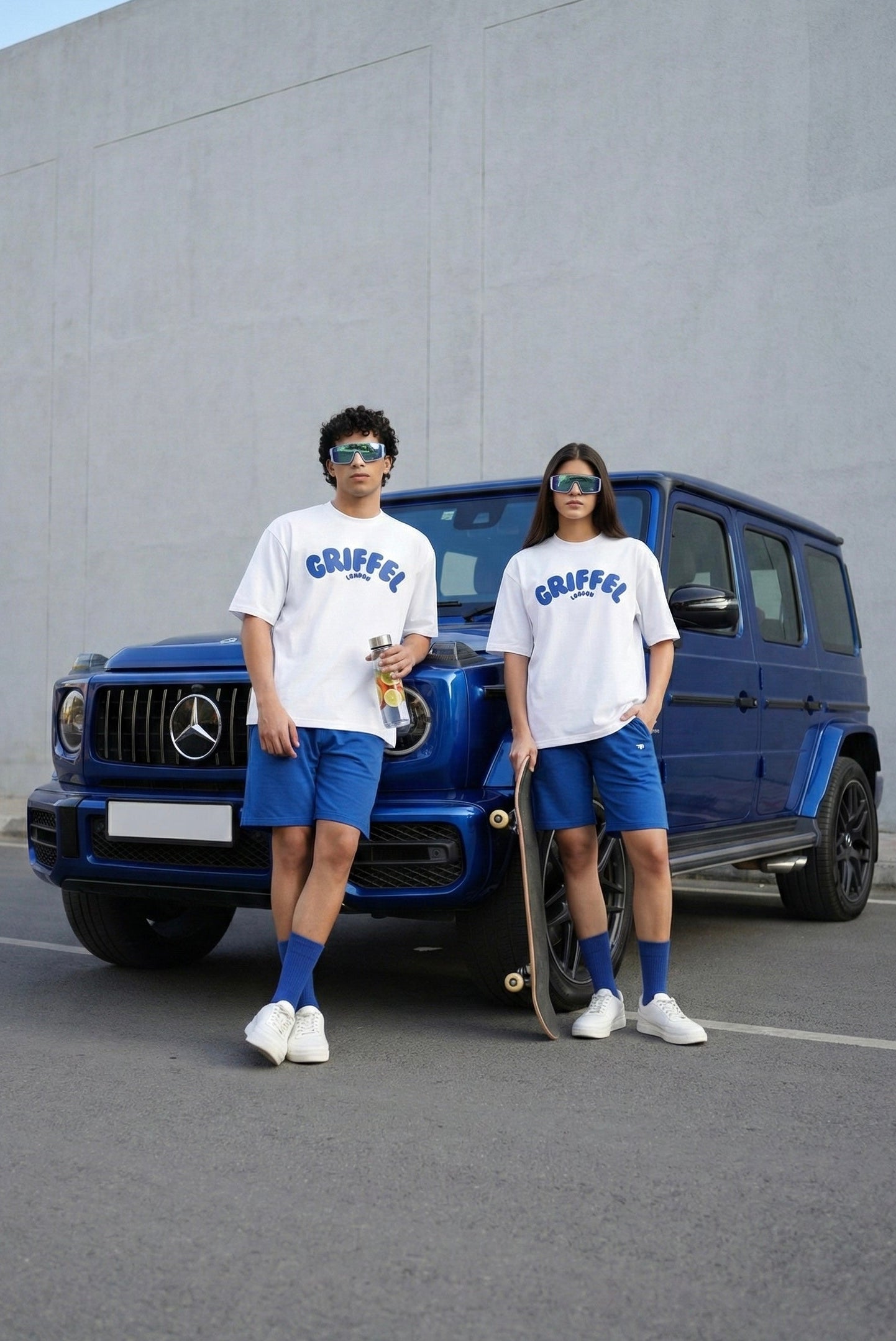 Two people in matching outfits standing next to a blue Mercedes-Benz G-Class SUV.