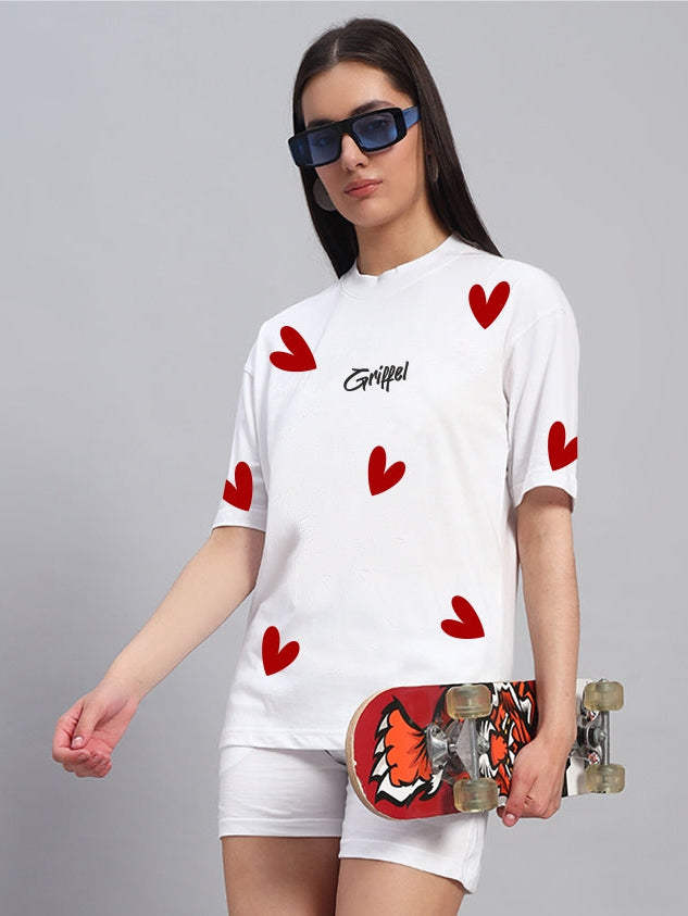 Woman holding a skateboard wearing a white t-shirt with red heart patterns and white shorts on a gray background